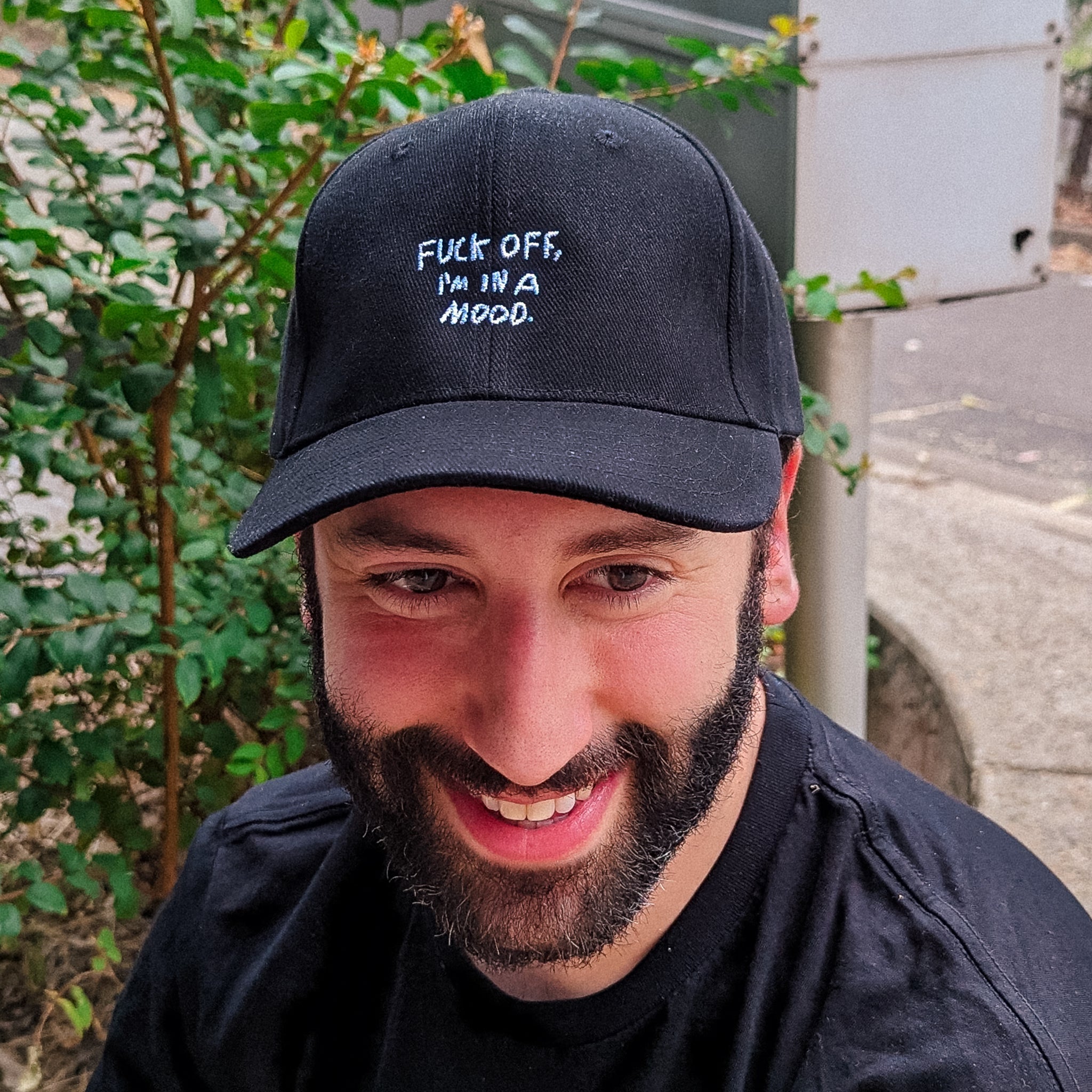 Bright outdoor close up photo of smiling man wearing a black cotton Fuck Off Cap by Lonely Kids Club.