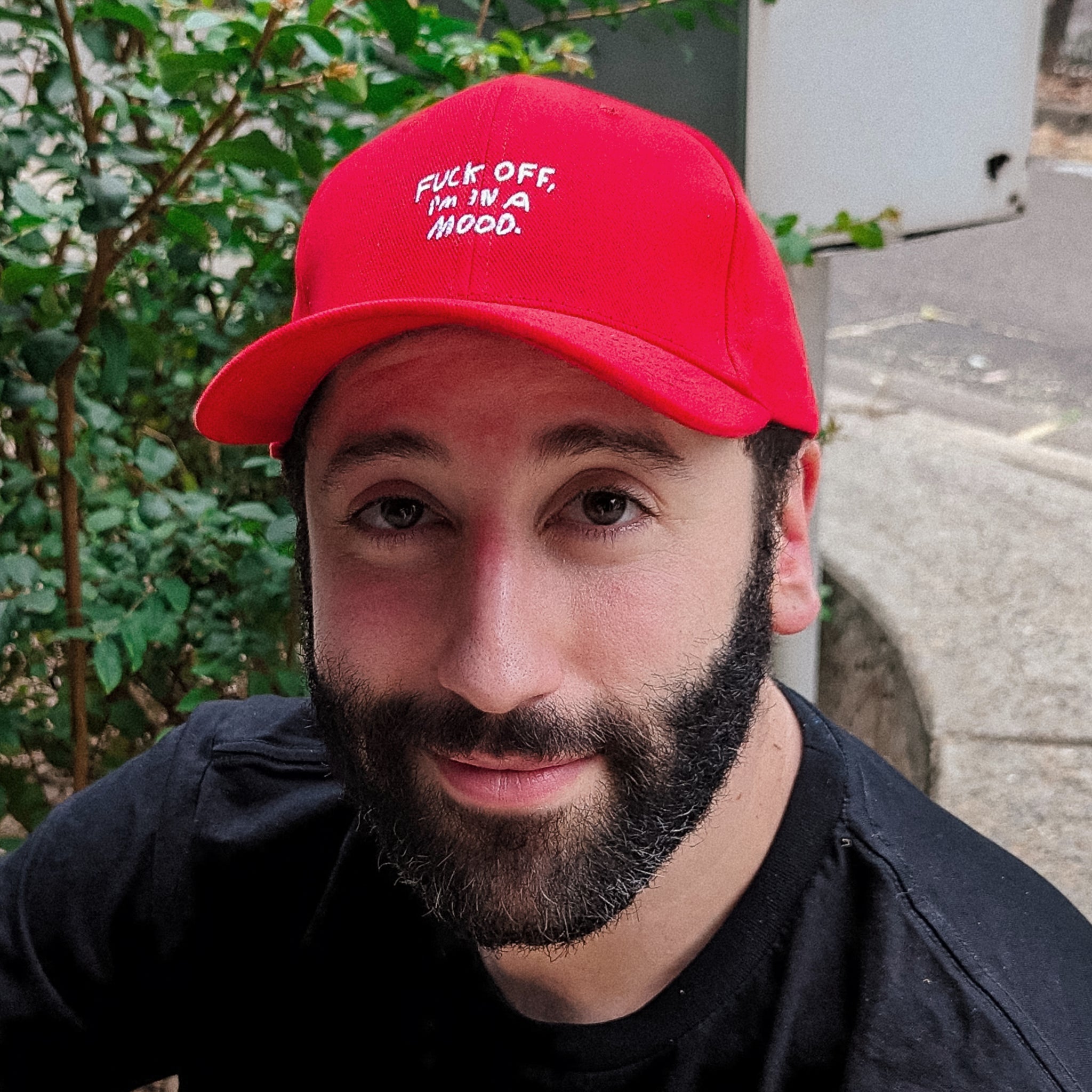 Close up photo with vintage effect of a bearded man happily wearing a bright red embroidered cap that says "Fuck off, I'm in a mood".